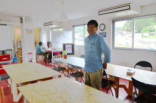 大きな窓から自然光が差し込む明るい室内で、複数のテーブルと椅子が配置され、中央に鈴木さんが立ち、奥のテーブルにも人が座っている室内の写真