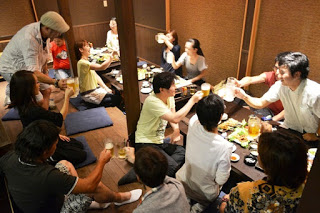 掘りごたつのテーブルが置かれた広い座敷で、大勢の人が2つのテーブルを囲み、飲み物を手に持って乾杯しているにぎやかな様子を写した写真