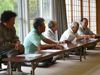 窓際に座った高齢の参加者たちがテーブルに肘をついて資料を見ながら真剣に話を聞いている写真