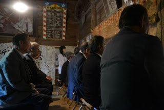 神社の内部でスーツ姿の男性たちが椅子に座って式典を見守っている写真