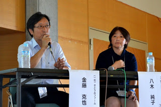 名札がつけられた長机の席に、男性と女性が並んで座り、左側の男性がマイクを持ちながら話をしている写真