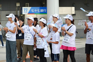 白い帽子とゼッケンをつけた白いTシャツを着た人々が屋外に集まって拍手をしたり旗を振っている写真