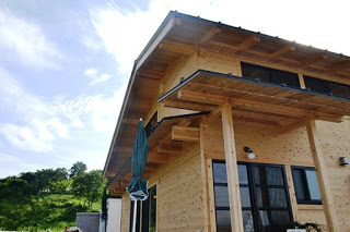 青空の下に広がる緑の丘を背景に、木造の家の一部が写っている写真