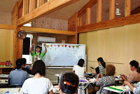 教室内のホワイトボードの前に女性講師が立ち、机に向かって座っている多くの参加者に対して手を上げて説明している写真