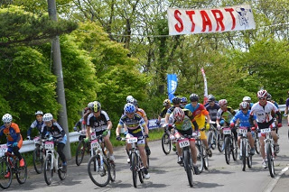 「START」と書かれた横断幕の下、ヘルメットとレーサーウェアを着た大勢のマウンテンバイク選手たちが一斉にスタートを切る瞬間の写真