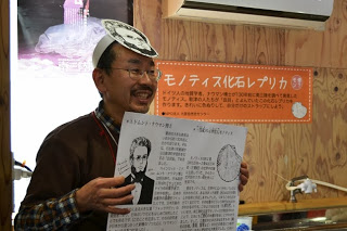 顔にハインリッヒ・エドムント・ナウマン博士が描かれたお面をかぶり、手に資料を持ちながら、来場者に笑顔で説明している男性スタッフの写真