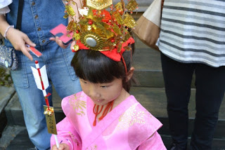 ピンクの着物を着て金色の冠をかぶった女の子が、赤と白の矢を手に持ち祭りの装いをした写真