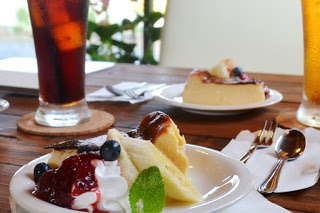 木製のテーブルの上に、ケーキと果物、生クリームが美しく盛られた白い平皿と、グラスに入った飲み物が並んでいる写真