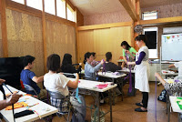 複数の参加者が机に向かって手作業をしており、女性講師とスタッフがそれぞれの席を回って指導している写真