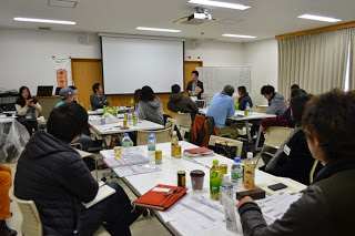 ホワイトボードとスクリーンのある明るい会議室で、前方に立つ男性の話を、複数のテーブルに分かれて座った参加者たちが話を聞いたりしている写真