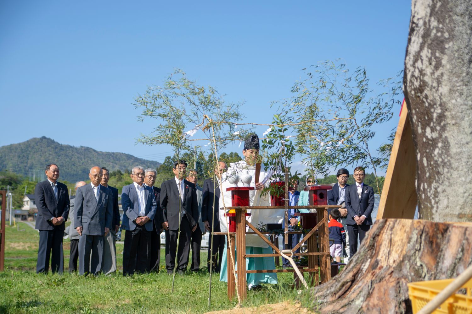 背景には木々と山々が広がる青空の下、大きな1本の木が立つ前で数人の人々が整列し、前方で白と水色の衣装を着た神主さんが神事を行っている写真