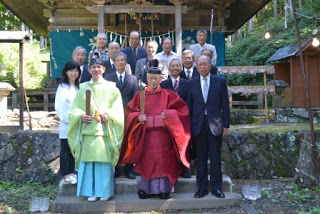神社の前で伝統衣装の宮司と神職の男性、参加者たちが記念撮影をしている集合写真