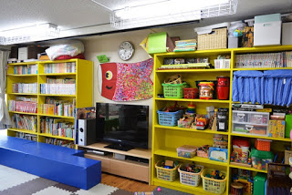 黄色い本棚に絵本が並び、ぬいぐるみやおもちゃがきれいに片付けられている、明るく整った室内の写真