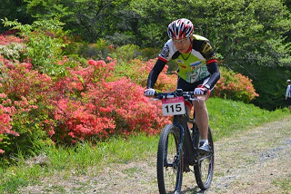 木々と赤いツツジの咲く山道を、ゼッケン115番の男性選手がを真剣な表情で走行しているレース中の写真