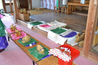 神社の拝殿前に色とりどりの衣装が広げられ、机の上には金色の冠や赤と白の矢が並べられている、祭りの準備道具が置かれた写真