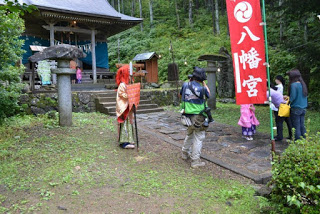 神社の参道に『八幡宮』と書かれた赤いのぼり旗が立ち、赤い髪ときらびやかな衣装をまとった人物やカメラを構える人々、着物姿の子どもたちが石畳の道を歩いている写真