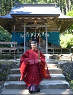 赤い伝統衣装を着て神社の階段に立つ宮司の男性の写真