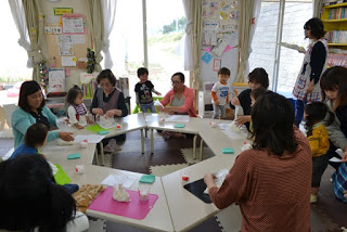 大きな窓から自然光が差し込む教室で、六角形に並べられた机のまわりに大人と子どもたちが集まり、活動に取り組んでいる様子の写真