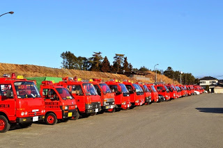 雲一つない青空の下、赤い消防団の小型消防車が十数台、整列して駐車場に並んでいる写真