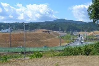 遠くに山々を望み、整備された道路の左にかさ上げ工事で造成された高台、右側に緑のフェンスが並ぶ風景の写真