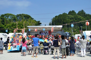 多くの人々が屋外のステージに集まり、演奏やパフォーマンスを楽しんでいる歌津復興夏まつりの様子の写真
