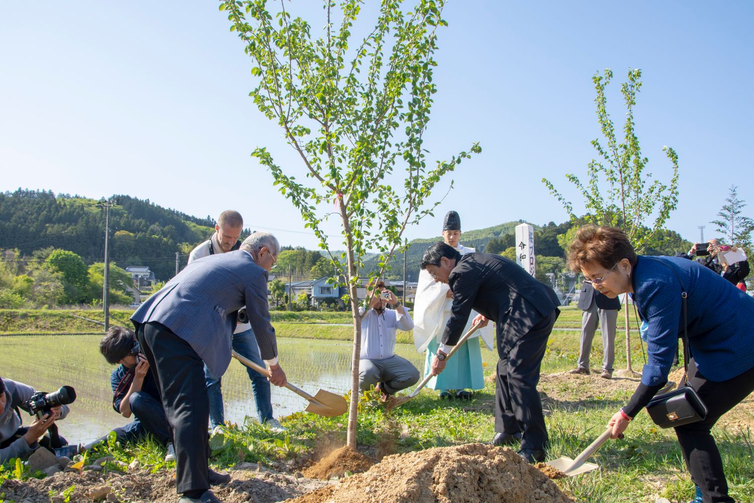 周囲には田畑や山、建物が点在し、周りでカメラマンが撮影するなか、数人の人々が若い木の根元に土をかけながら植樹活動を行っている写真