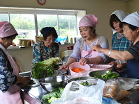 調理室で、エプロンと三角巾をつけた女性たちが野菜を切ったり洗ったりして料理の準備をしている様子の写真