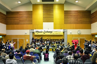 黒い衣装を着た吹奏楽団が演奏を披露し、客席には多くの観客が静かに耳を傾け、ステージの右側には赤いマスコットキャラクター「チーバくん」が見える会場内の写真