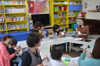 本棚やカラフルな収納ボックスが並ぶ室内で、大人と子どもたちが机を囲んで座り、かんたんクッキー作りに取り組んでいる様子の写真