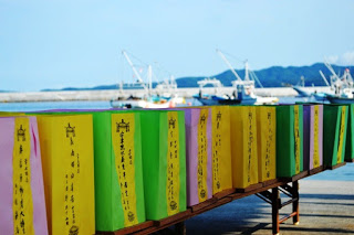 港を背景にしてピンク、黄色、緑の色とりどりの灯ろうが長机に並べられている写真