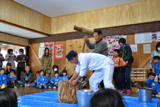 ブルーシートが敷かれた室内で手前の白い服の人がもちをこね、後ろで大きな木槌を振りかぶっている人が餅つきをしている様子を青いスモックを着た子どもたちと、保護者・スタッフと思われる大人たちが取り囲んで見学している写真