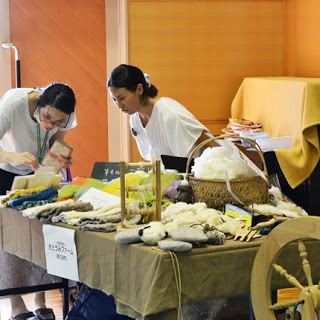 手作りの毛糸や布製品が並べられたテーブルの前で女性二人が作品を覗き込んでいる写真