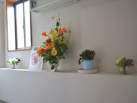 左上の窓から光が差し込み、白い壁の棚の上に、オレンジや黄色の花が入った大きな花瓶を中心に、小さな鉢植えや花が生けられた透明の小さな花瓶や観葉植物が並んでいる写真