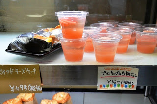 冷蔵ショーケースに並ぶ透明カップ入り白桃のゼリーと、隣にチーズケーキが並び、それぞれに手書き風の価格札が付いているスイーツの写真