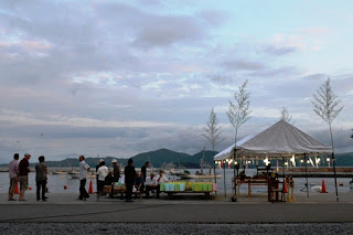 夕暮れ時の漁港にライトが付いた簡易的なテントが設置されており、その周囲に人々が集まっている写真