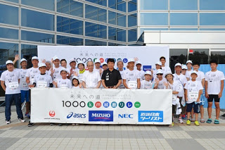 白いTシャツを着た大勢の人々が建物の前に集まり、「未来への道 1000キロメートル縦断リレー」と書かれた横断幕を広げている写真