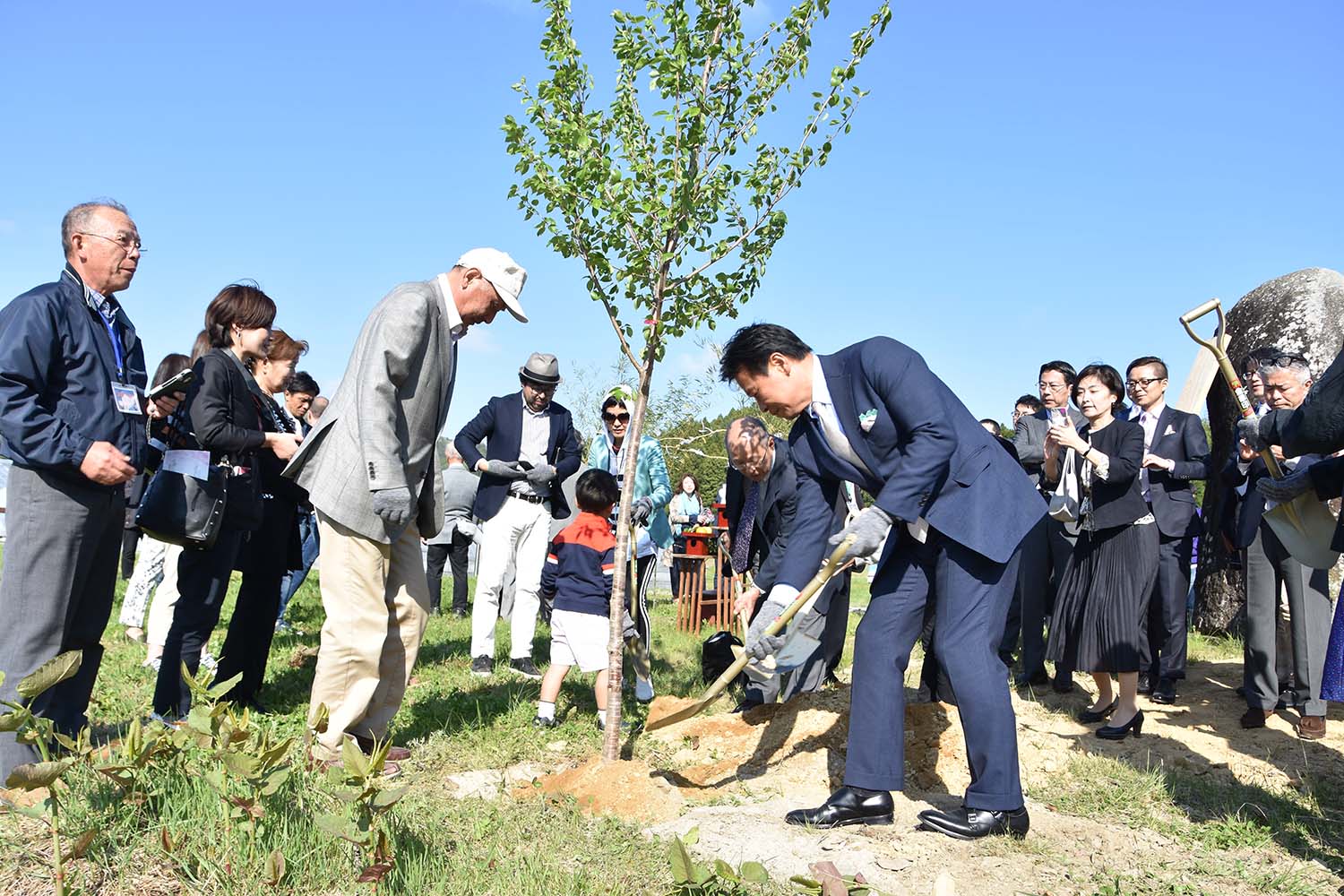 フォーマルな服装をしている参加者が写真を撮ったり見守ったりしているなか、若い木の周りに数人の人々が集まり、そのうちの一人が木の根元に土をかけている植樹の様子の写真