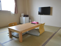 壁にテレビが掛けられ、小型の冷蔵庫や電気ポットが置かれている部屋の中央に、木のテーブルと座椅子が置かれた和室の写真