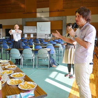 ジャムを乗せたクラッカーがたくさん盛られた皿が複数並んだテーブルの前に立ち、マイクを持って話している男性と、それを囲んで見ている数人の様子が写っている写真