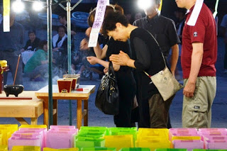 灯ろうが並んだテントの下で数名の人が手を合わせて焼香をしている様子を写した写真