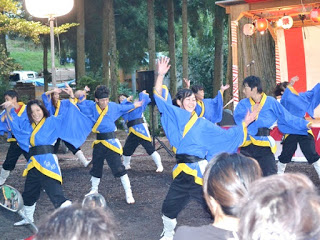 青と黒の衣装の若者たちが笑顔で元気よく踊っており、背景には木々と提灯で飾られた舞台が見える写真