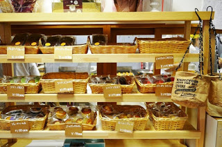 木製の棚に並んだ十数種類の焼き菓子が種類ごとにカゴに入れられ、それぞれの前に手書き風の値札が付けられている陳列棚の写真