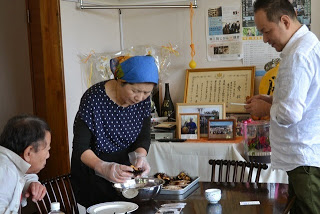 女性が室内の食卓で料理を盛り付けており、二人の男性がそばで料理に注目していて、後ろの棚には賞状や写真が飾られている写真