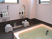 浴槽にお湯が張られ、洗い場にはシャンプー類がセットされたシャワーが2つ並び、腰かけ用の椅子と洗面器が置かれている浴室の写真