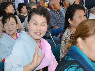 和装をした年配の女性が観客席から笑顔で手を振っており、その周囲には同年代の観客が多数座っている写真