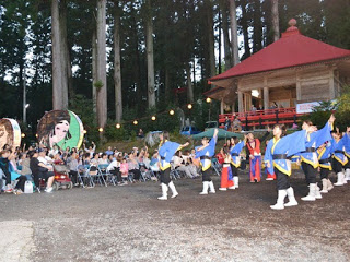 神社境内の舞台の前で、青い法被を着た踊り手たちが整列して手を上げ、観客が拍手している写真