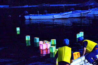 黄色いライフジャケットを着た二人の男性が小舟から次々と灯ろうを海に流し、暗い水面に色とりどりの灯ろうが静かに浮かんでいる写真