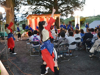 赤や青のカラフルな衣装を着たダンサーが、ステージ前で両手を大きく上げながら踊っている様子を観客が見守っている写真