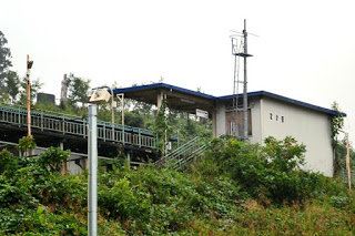 小さな無人駅のホームと待合室が草木が生い茂った斜面の上に建っており、ホームの左側に四角い石碑が見える写真