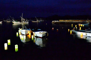 夜の港に小舟が静かに浮かび、その周囲には灯ろうが浮かぶ幻想的な風景の写真